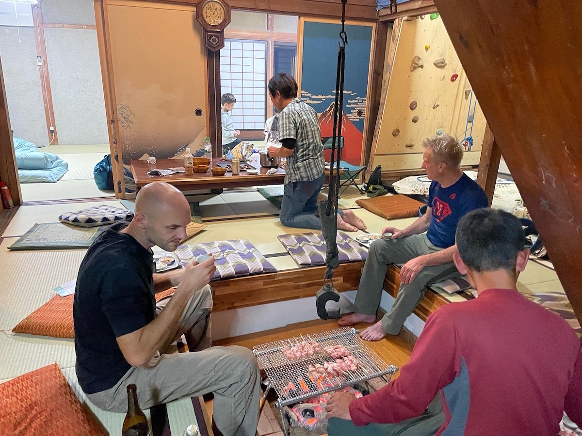 A cozy indoor space is set up for a barbecue gathering, featuring a low wooden table surrounded by cushions. Four individuals are seated and enjoying grilled food, with a traditional Japanese room visible in the background, showcasing tatami flooring and shoji screens.