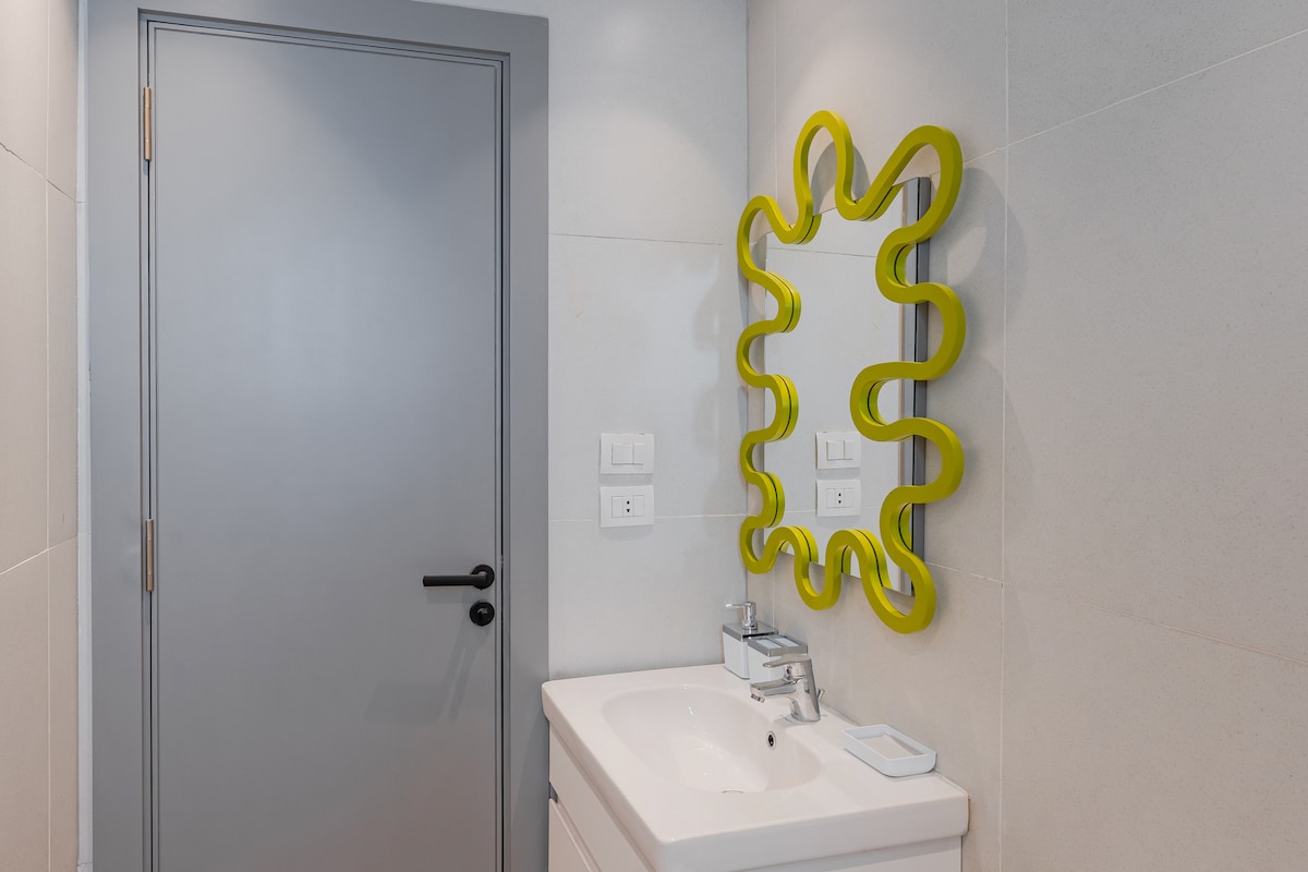A modern bathroom features a white sink counter with a sleek faucet and minimal accessories. A uniquely shaped yellow mirror enhances the space, reflecting light. Light-colored tiles create a clean environment, and a gray door is positioned in the background.
