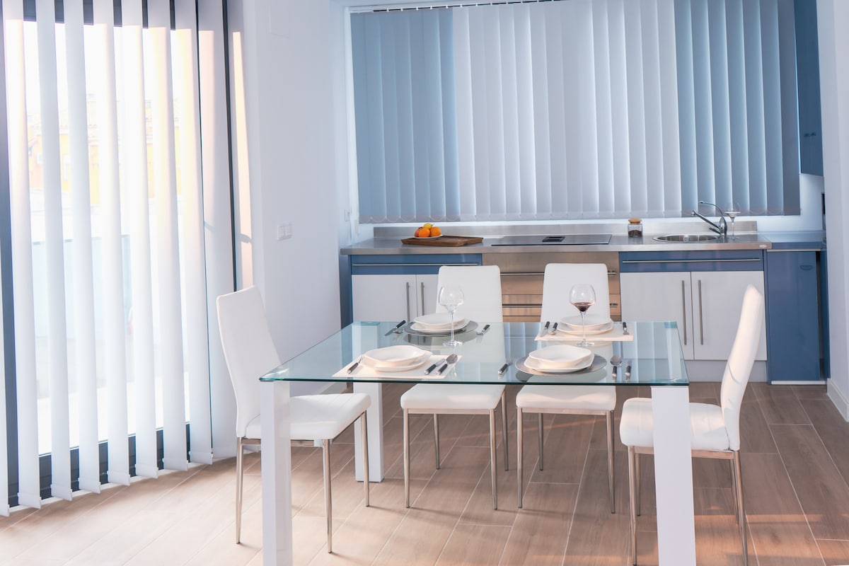A modern kitchen area is presented with a glass dining table surrounded by four white chairs. Light pours in through vertical blinds, illuminating the space. The kitchen features sleek cabinets and an organized countertop with essential appliances visible.