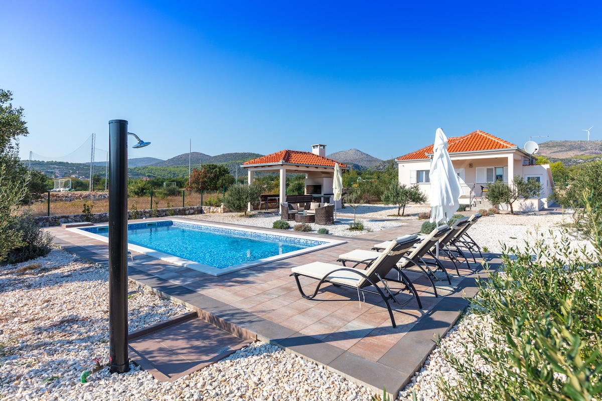 A large swimming pool is surrounded by sun loungers, inviting relaxation under the clear blue sky. A villa with a red-tiled roof and a shaded outdoor dining area can be seen in the background, nestled among olive trees and gravel pathways.