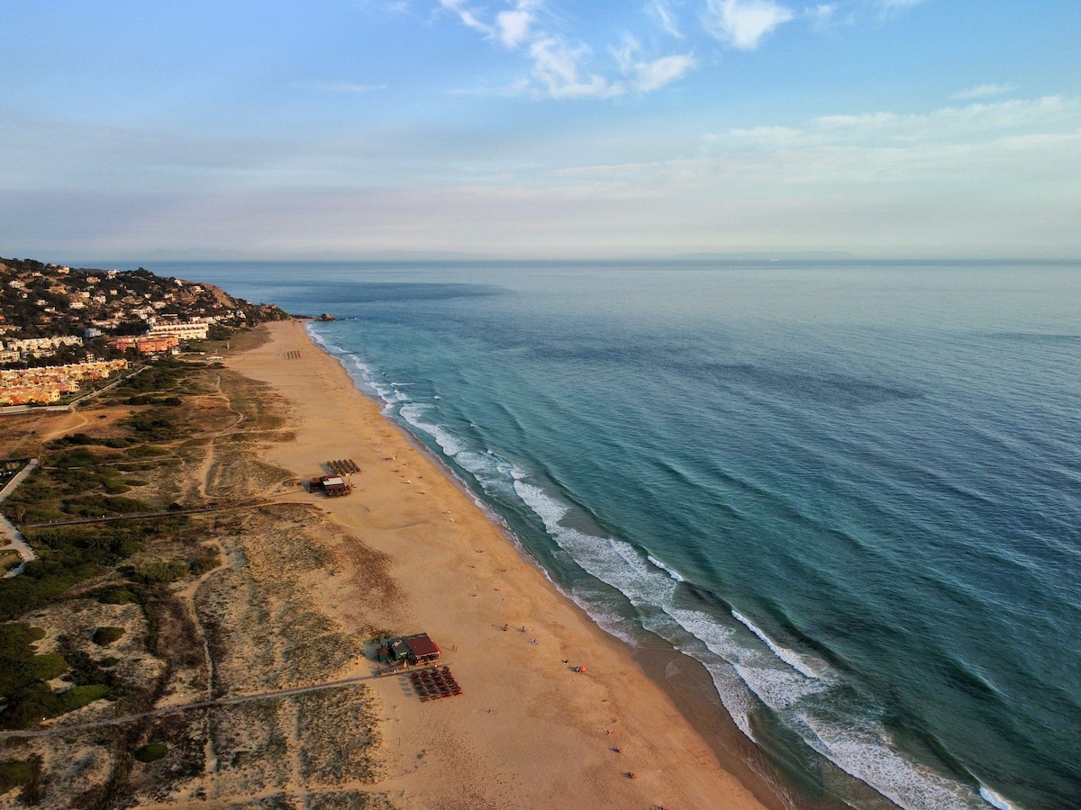A sweeping view of a sandy beach stretches along the shoreline, where gentle waves meet the shore. Coastal vegetation is visible alongside several beach umbrellas and lounge chairs positioned on the sand. The horizon showcases a serene blend of sky and sea.