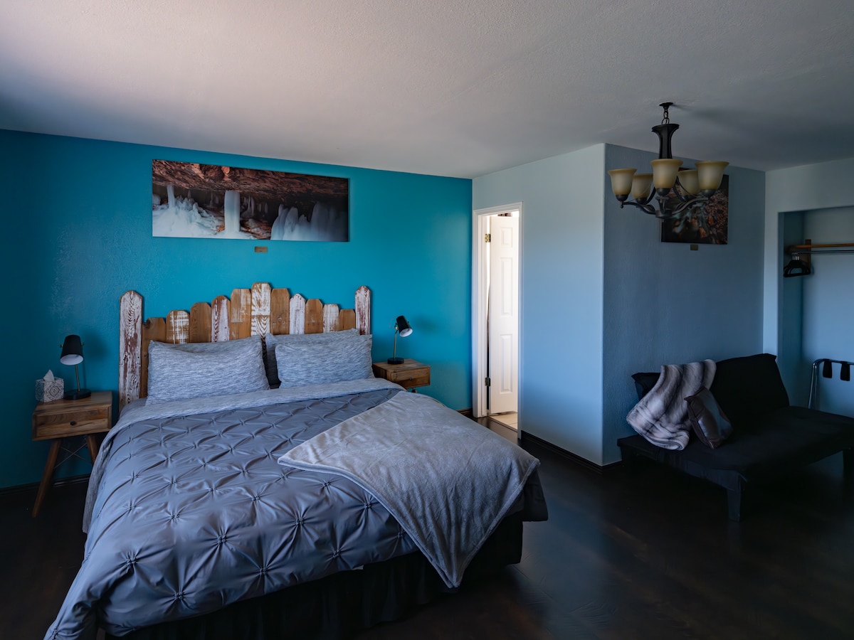 A spacious sleeping area features a large bed with a textured gray comforter and two bedside tables with lamps. The vibrant blue wall adds color, while a portrait hangs above the bed. A small sofa is positioned nearby, and a doorway leads to another space.