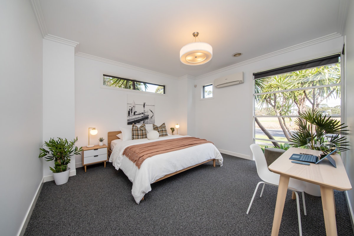 A spacious bedroom features a large bed with neutral bedding and decorative pillows. Natural light is welcomed through two windows that frame views of greenery. A small desk with a chair and potted plants complement the room's decor, creating a relaxed atmosphere.