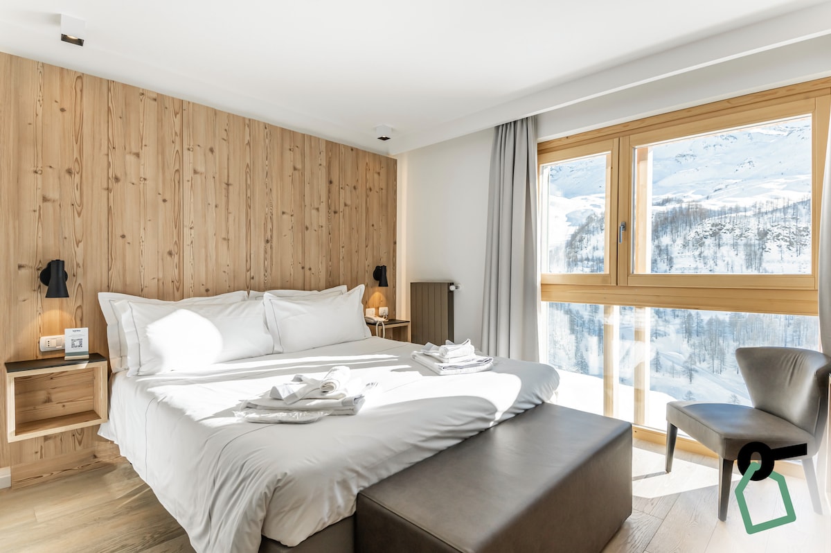 A spacious bedroom is featured, characterized by wooden accent walls and large windows showcasing mountain views. A comfortable bed is dressed in white linens, accompanied by neatly arranged towels. A cozy seating area is provided by a dark ottoman near the foot of the bed.