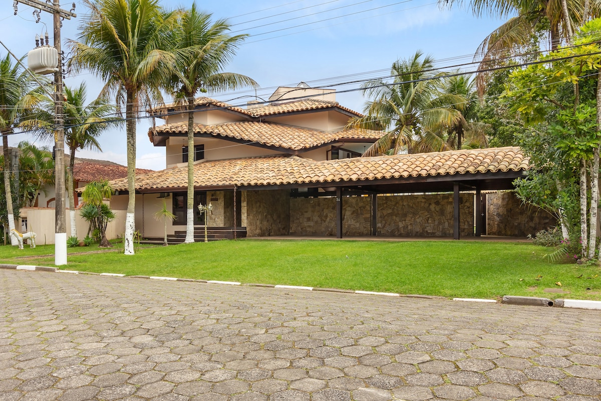 A two-story house with a sloped tiled roof is set in a lush green landscape. Palm trees provide shade in the front yard. A spacious driveway and a stone wall feature can be seen, contributing to the welcoming exterior.
