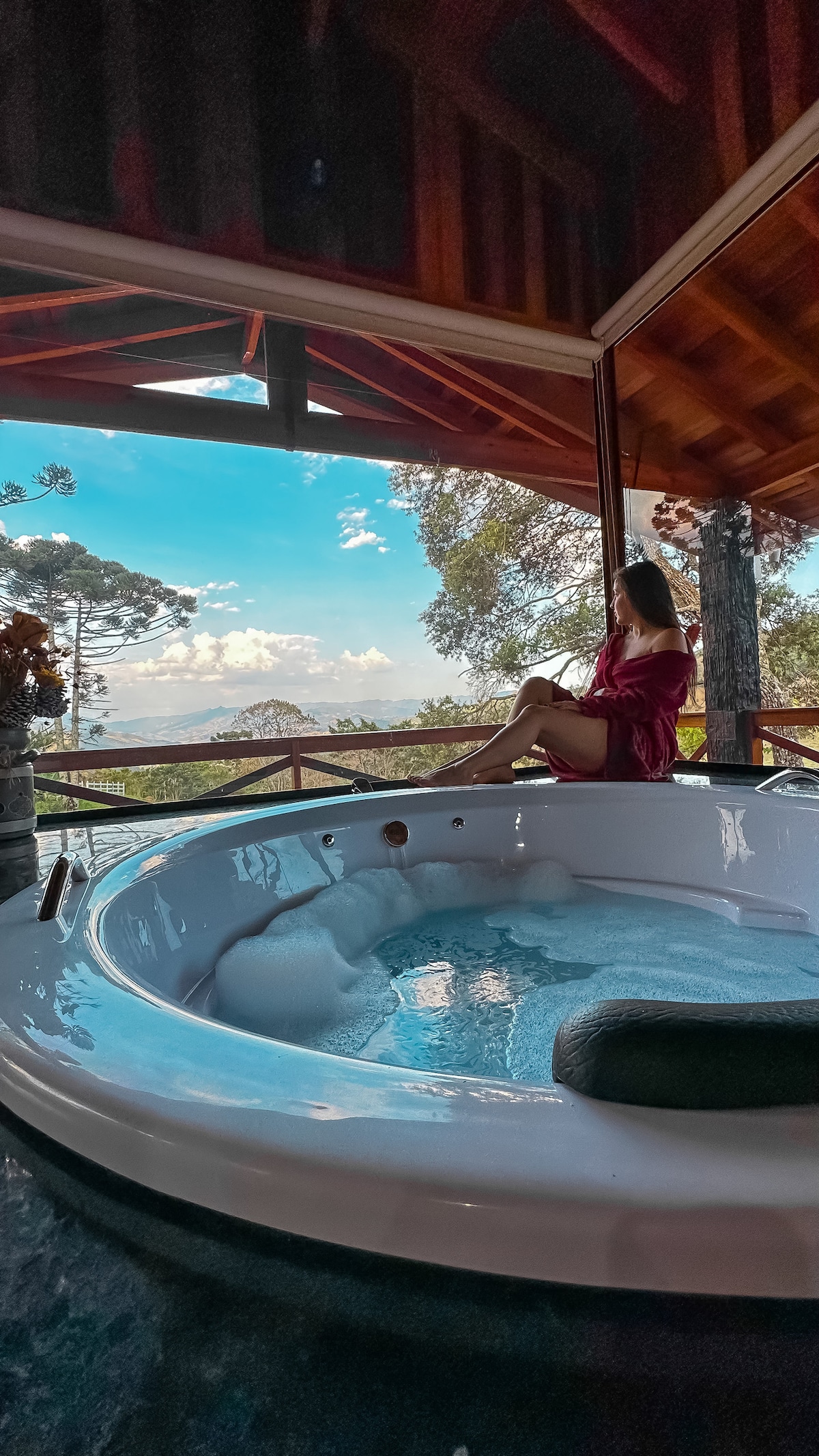 A spacious hot tub is positioned next to large glass doors, offering a stunning view of the surrounding landscape. Natural light floods the area, highlighting the serene atmosphere. The rich wooden ceiling contributes to the cozy cabin ambiance.
