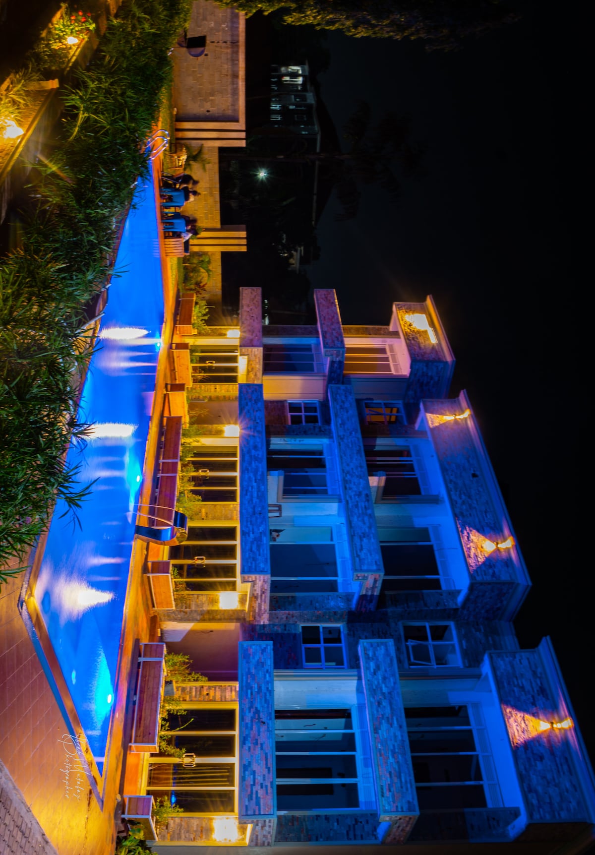 A modern building is showcased at night, featuring large windows and illuminated by warm outdoor lighting. A swimming pool with a vibrant blue hue reflects the lights, surrounded by benches and lush greenery. Guests can be seen enjoying the space in the background.