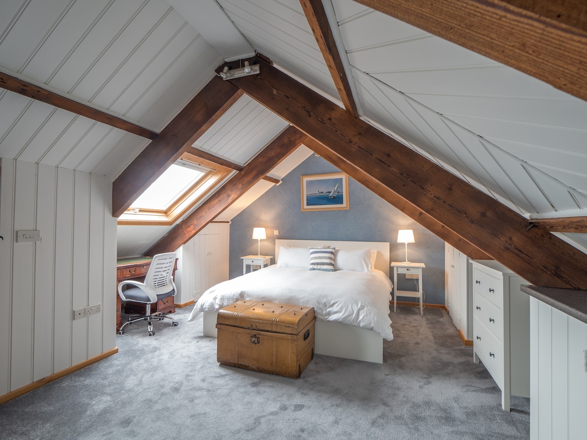 An inviting attic bedroom features a spacious bed centered under angled ceilings, complemented by soft carpeting. A wooden trunk and a chair with a desk are positioned to one side, while warm light is provided by bedside lamps. Large skylights allow natural light to fill the room.