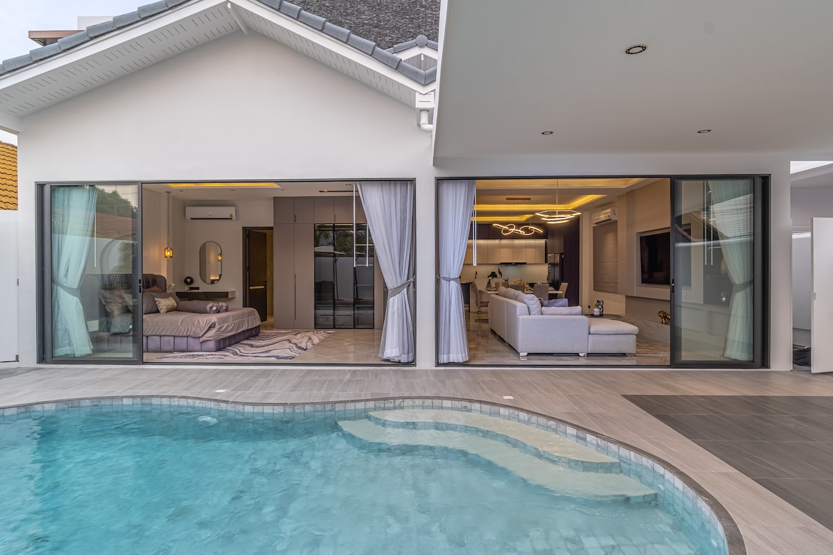 A modern villa is shown, featuring large glass doors that provide an open view of both the bedroom and living area. The inviting private pool reflects the surrounding architecture, creating a seamless connection between indoor and outdoor spaces.