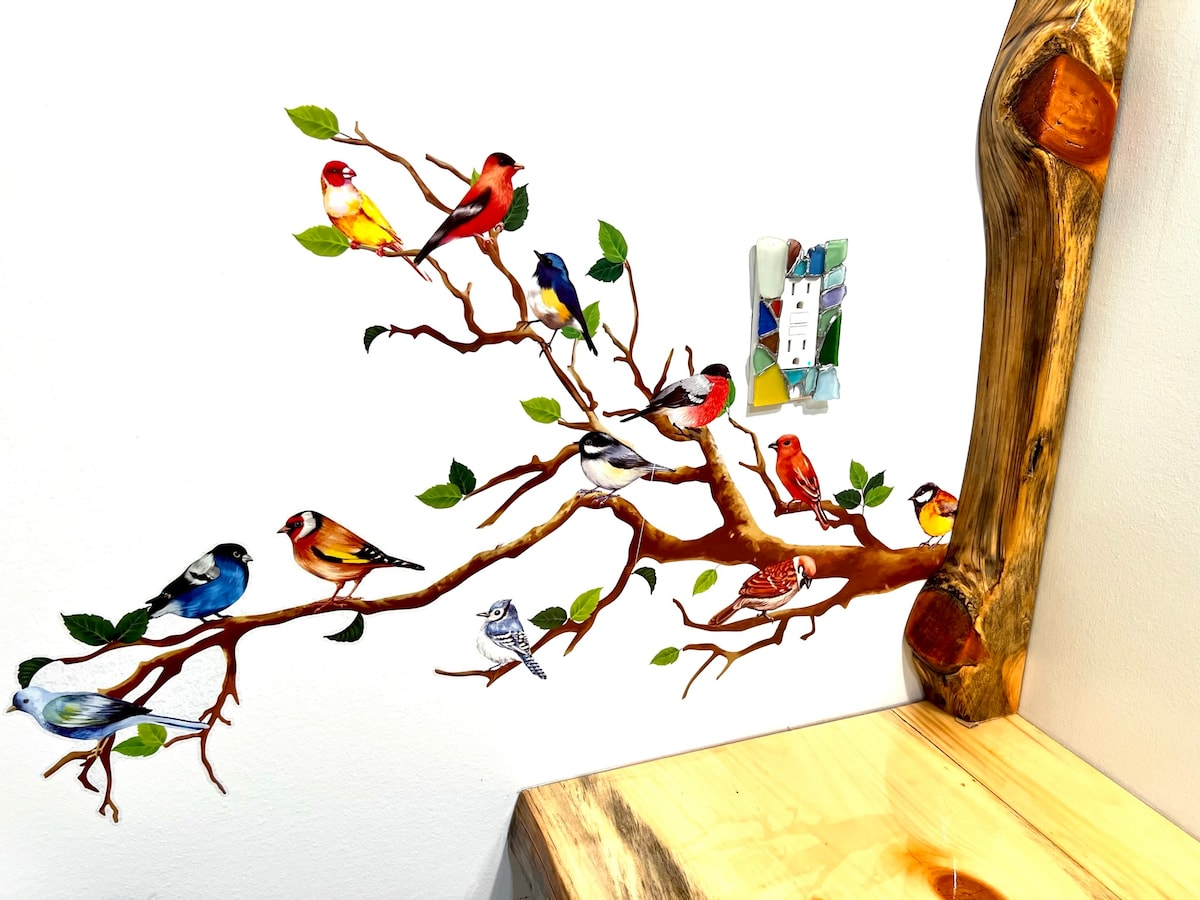 A mural depicting colorful birds perched on a painted branch enhances the wall. Various species are illustrated, showcasing their vibrant plumage among green leaves. The rustic wooden edge of a nearby table complements the natural theme.