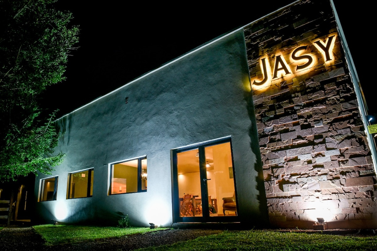 A modern building exterior is illuminated at night, featuring large windows that allow glimpses of the interior. The name 'JASY' is prominently displayed in glowing letters above a textured stone wall, enhancing the contemporary design. Soft landscaping elements are visible in the foreground.
