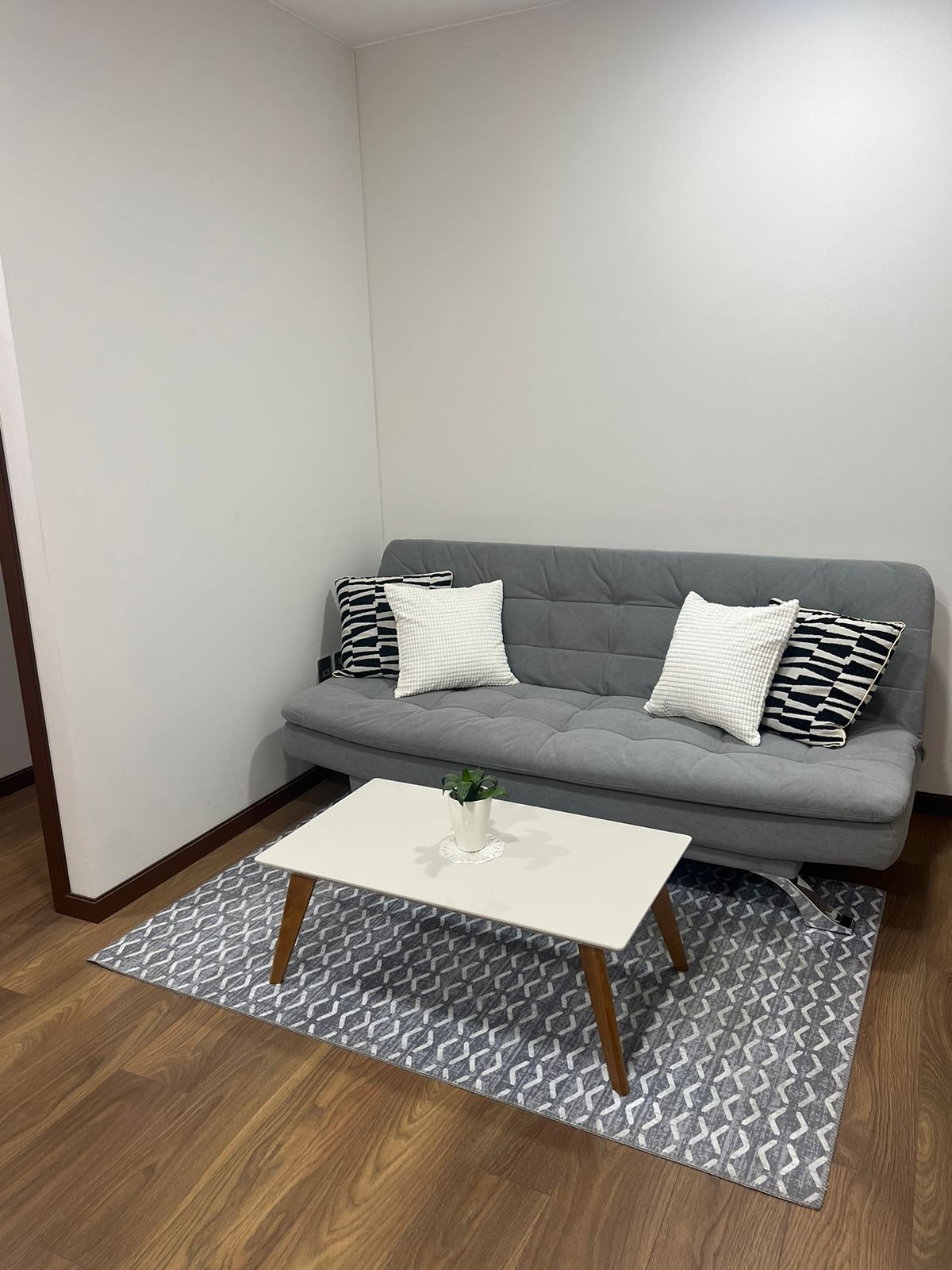 A cozy sitting area is highlighted by a gray futon adorned with striped and patterned cushions. A small white coffee table is positioned in front, resting on a textured area rug. The wall is painted in a subtle light color, complementing the warm wood flooring.