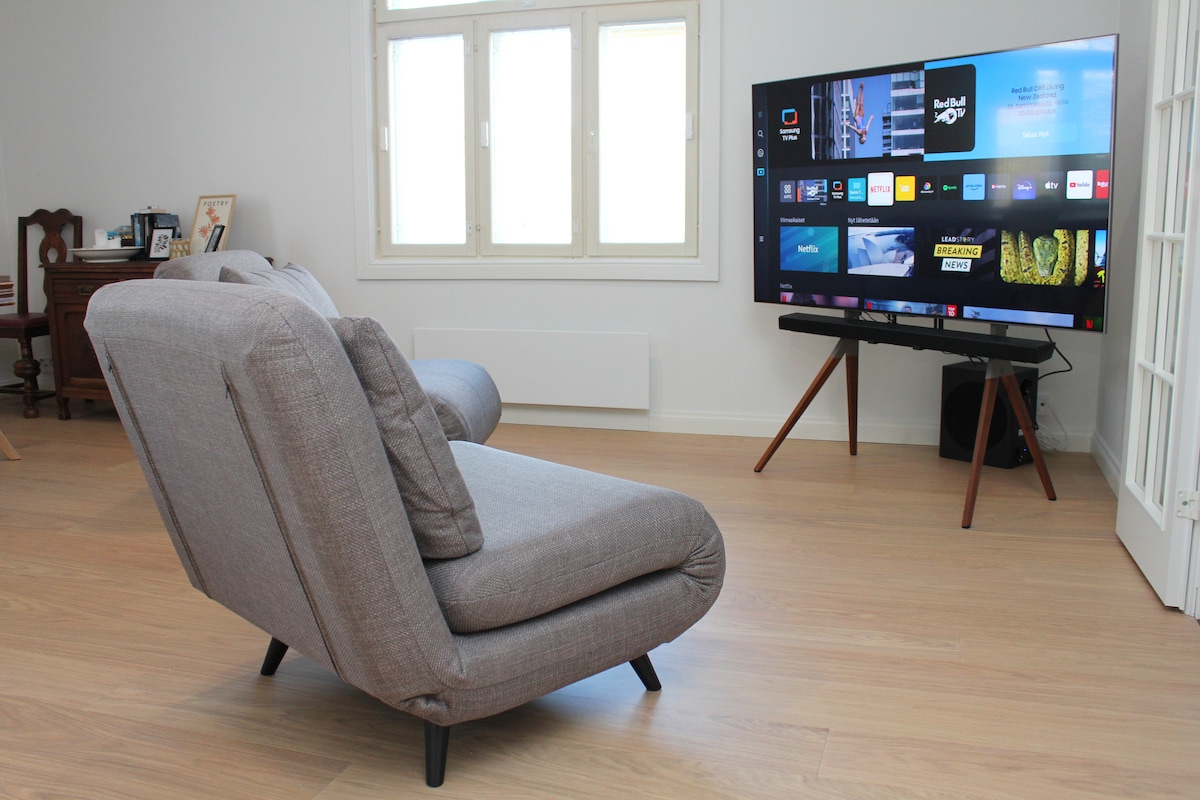 A comfortable, grey accent chair is positioned in front of a large flat-screen television mounted on a stand. Natural light fills the room through several windows, enhancing the relaxed ambiance of the space. A minimalist design is evident in the room's decor.