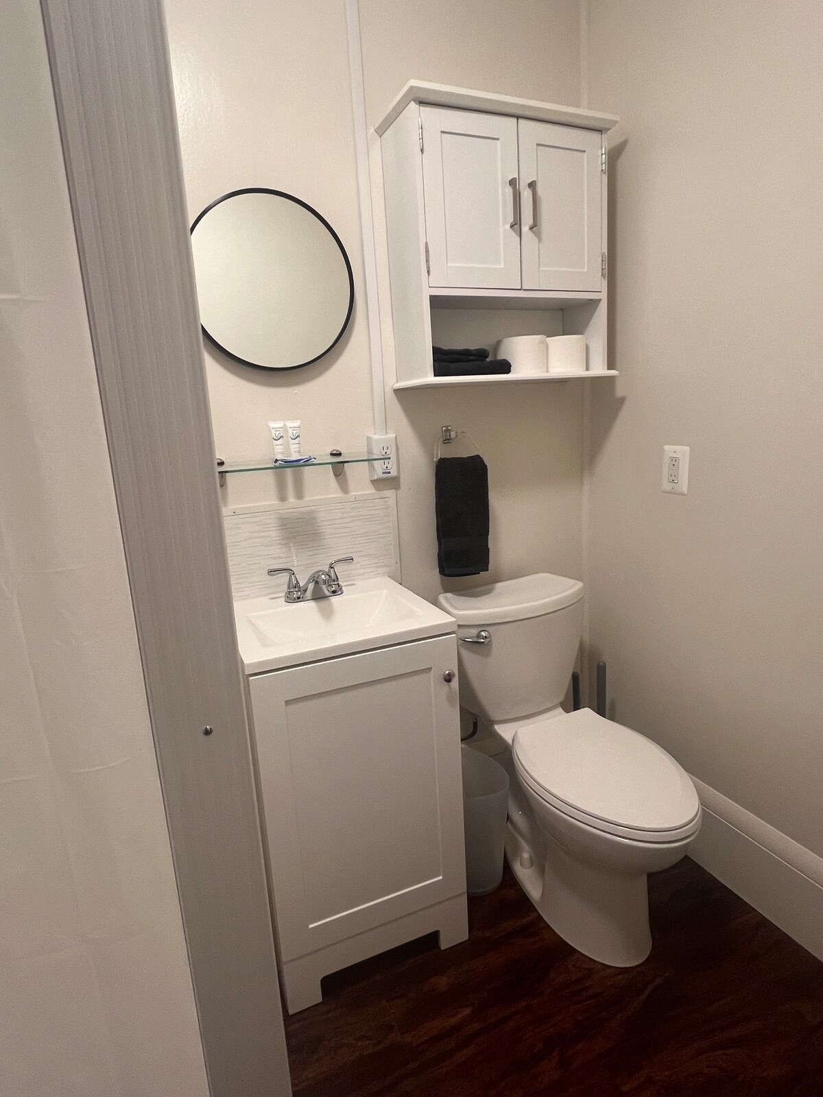 A compact bathroom features essential amenities including a white toilet and a small sink with modern fixtures. Above the sink, a shelf holds neatly arranged towels and toiletries. A round mirror adds depth to the space, complemented by light-colored walls and dark wood flooring.