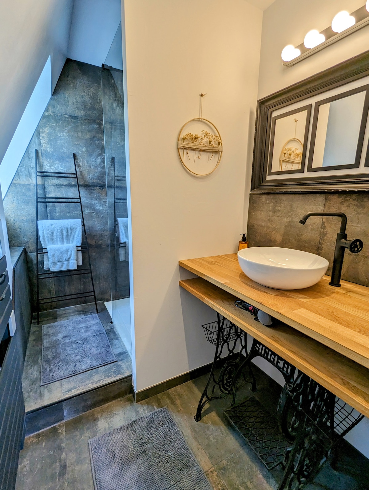 The bathroom features a modern design with a walk-in shower and decorative touches. A round basin is positioned on a wooden countertop, complemented by a vintage-style table base. Soft towels hang on a black ladder drying rack, and elegant lighting enhances the room's charm.