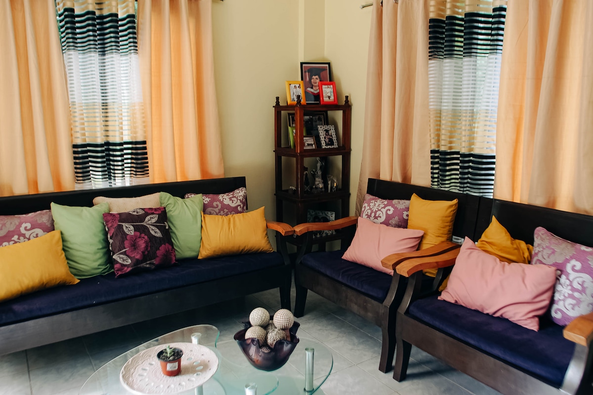 A comfortable seating area is highlighted by dark wooden furniture with plush cushions in various colors. Light-colored curtains frame the windows, allowing natural light to brighten the space. A glass coffee table sits centrally, complemented by decorative items such as a bowl of ornamental spheres.