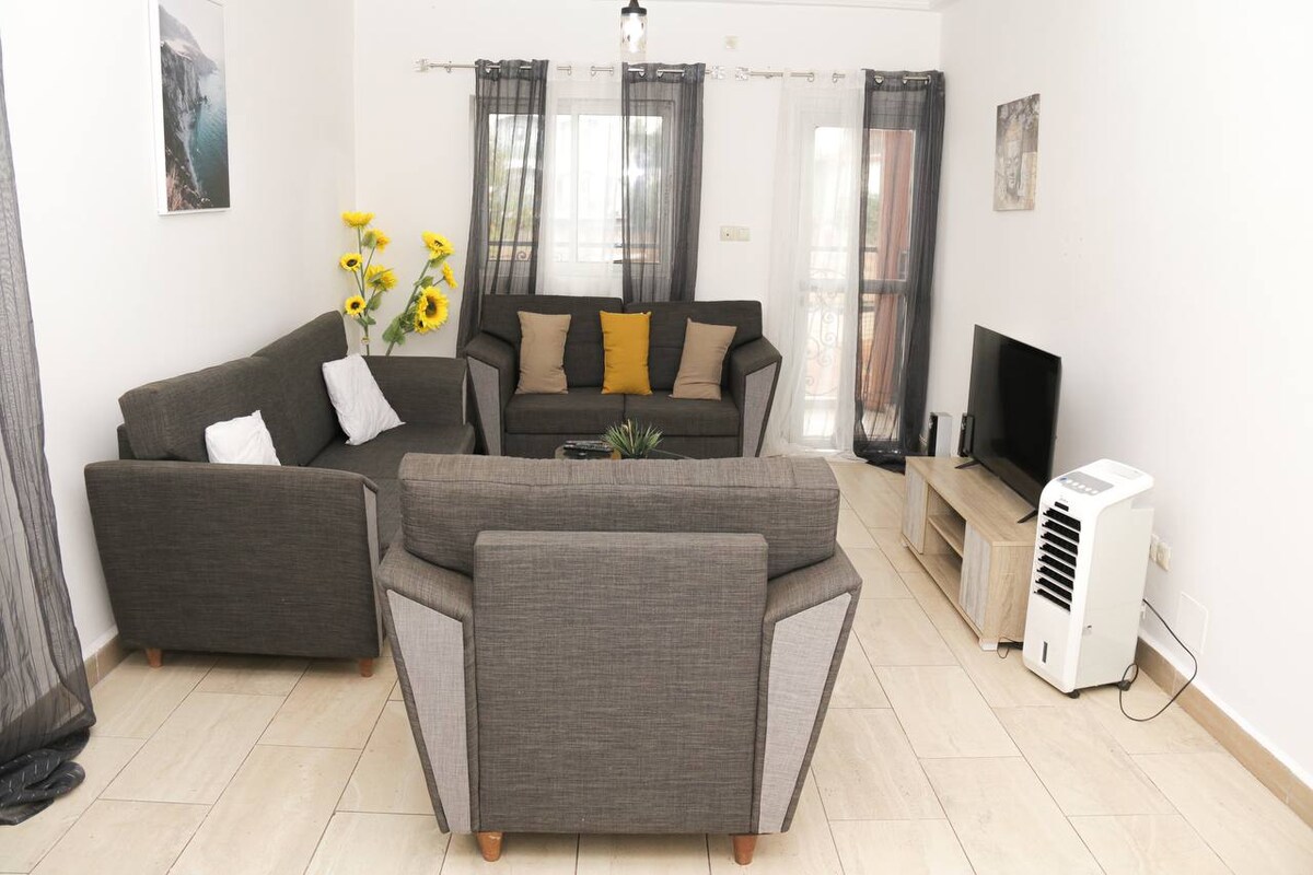 A spacious living area is furnished with a gray sectional sofa and an additional gray armchair, accented by yellow and gray pillows. Natural light filters through sheer curtains, illuminating a television on a wooden stand, with a decorative plant visible on the coffee table.