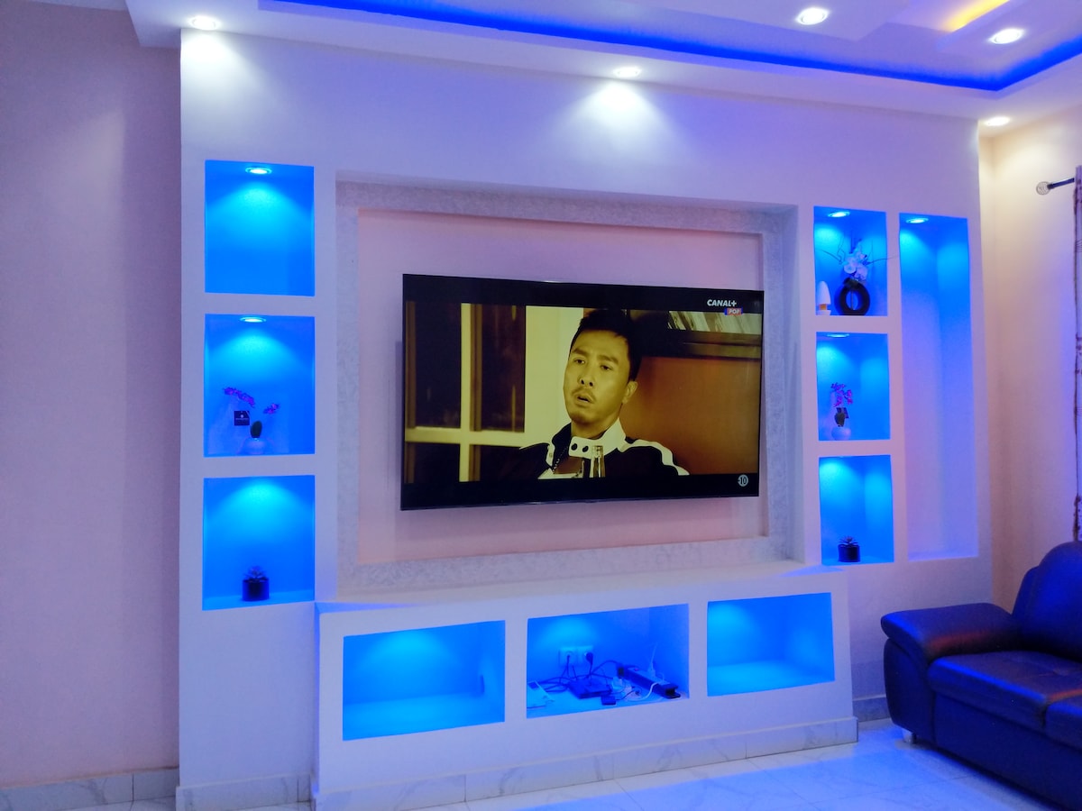 A modern living area features a large wall-mounted television framed by illuminated shelves in calming blue tones. Decorative items are displayed on the shelves, and a dark leather sofa is positioned nearby, creating a comfortable environment for relaxation.