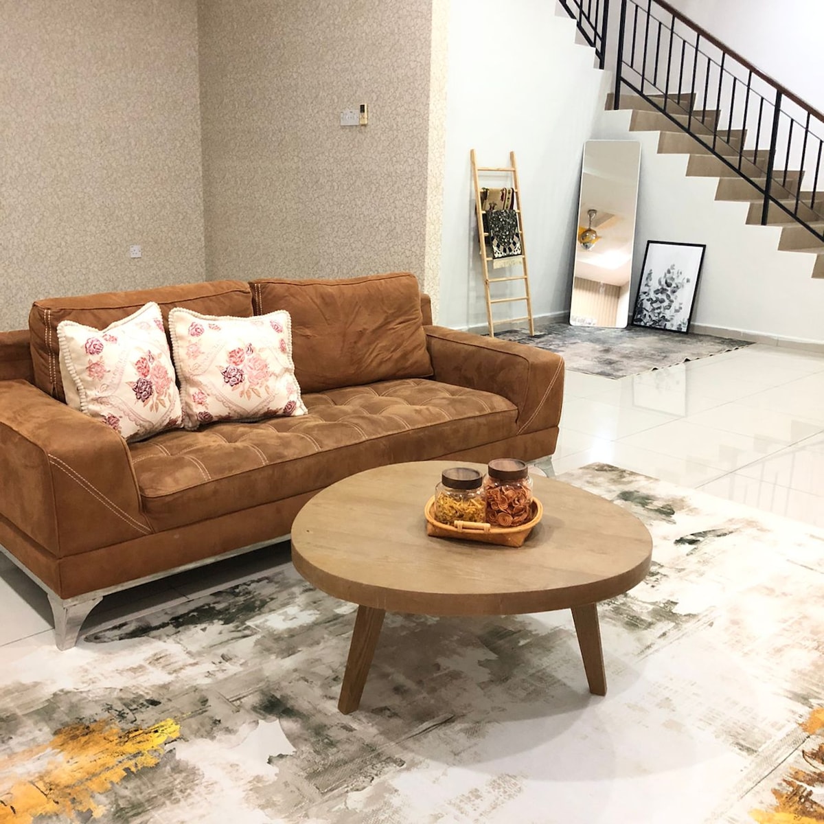 A cozy living area features a brown sofa adorned with floral-patterned cushions. A round wooden coffee table is arranged with snacks. Light-colored flooring complements the neutral walls, and a decorative mirror reflects the space. A staircase is visible in the background.