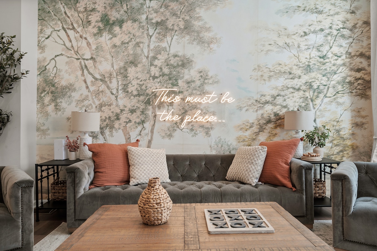 A stylish living area features a comfortable grey sectional sofa adorned with soft cushions in warm tones. An elegant coffee table made of natural wood sits at the center. Behind the sofa, a vibrant wall mural depicting serene trees creates a relaxing ambiance, complemented by two tasteful lamps.