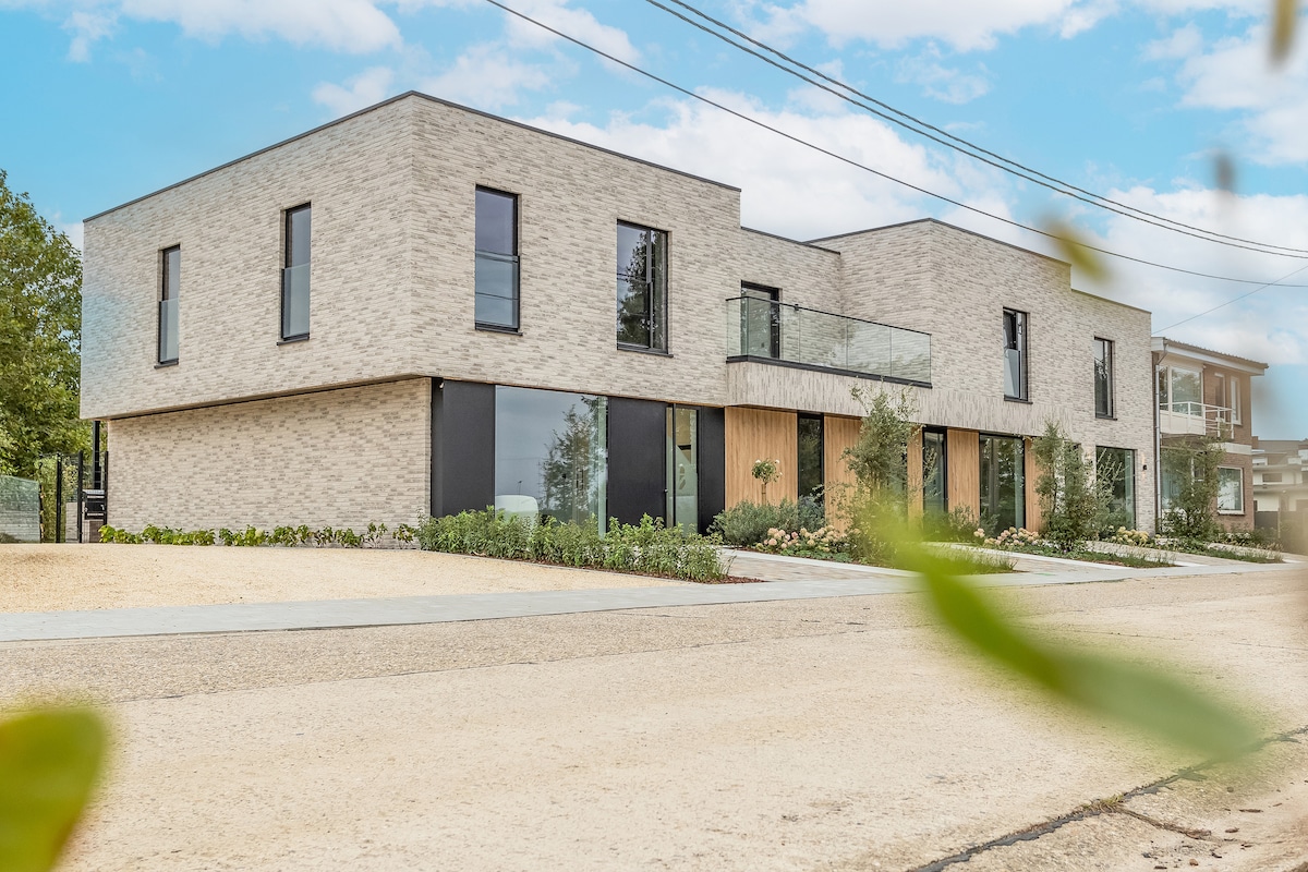 The modern building, characterized by clean lines and a neutral brick facade, features large windows that allow ample natural light. The entrance is accented by wooden elements, while landscaped greenery surrounds the property, offering a welcoming aspect to the exterior.