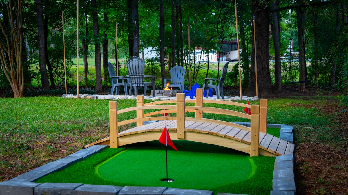 A mini-golf area features a green putting surface framed by landscaping, with a wooden bridge crossing over. In the background, comfortable seating arrangements surround a fire pit, set against a backdrop of trees. Red flags mark the putting cup.