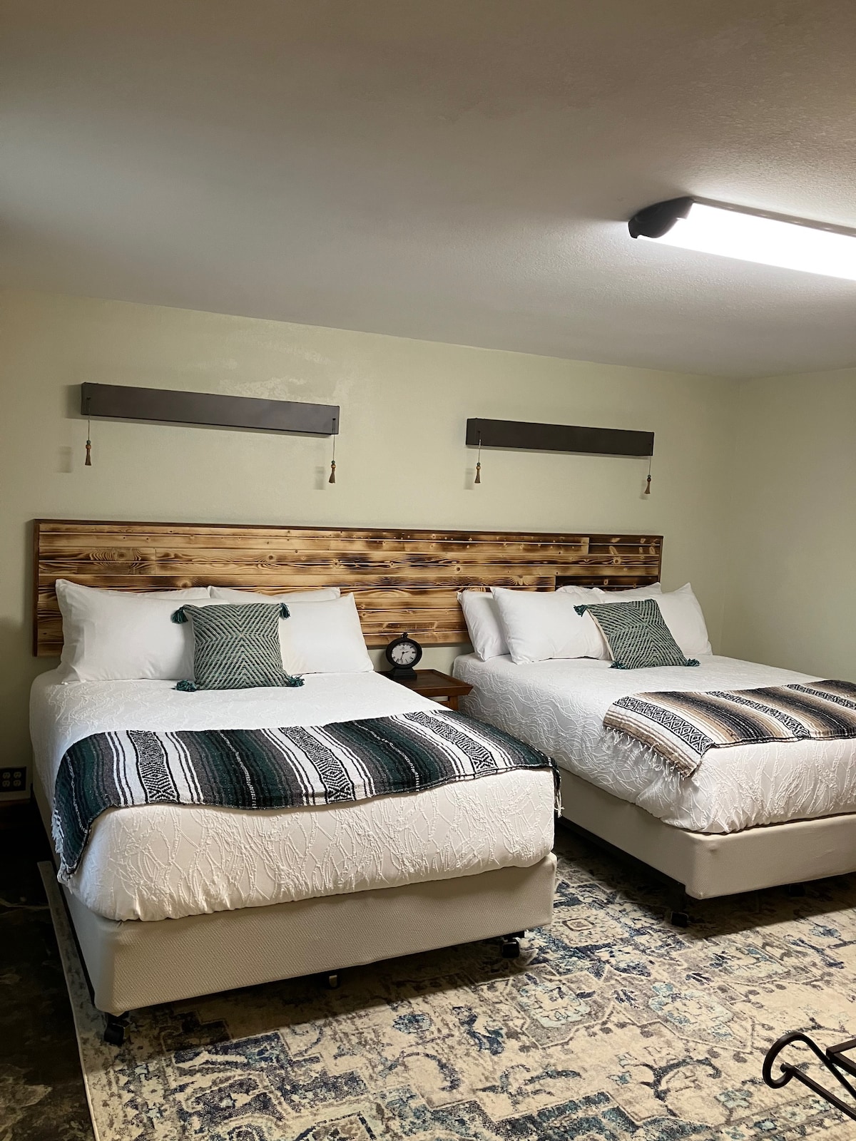 A bright bedroom features two spacious beds with patterned blankets and decorative pillows, set against a wooden headboard. Soft lighting from overhead enhances the welcoming atmosphere. A decorative rug covers the floor, while a small clock is positioned on a bedside table.
