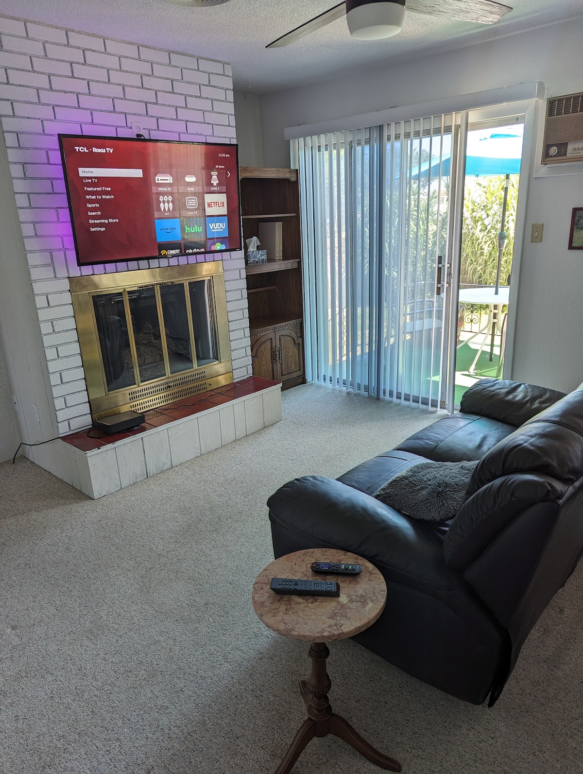 A comfortable living area features a black leather sofa and a small side table with remote controls. A wall-mounted television is positioned above a white brick fireplace. Light streams in through sliding glass doors, which lead to an outdoor space with patio furniture.
