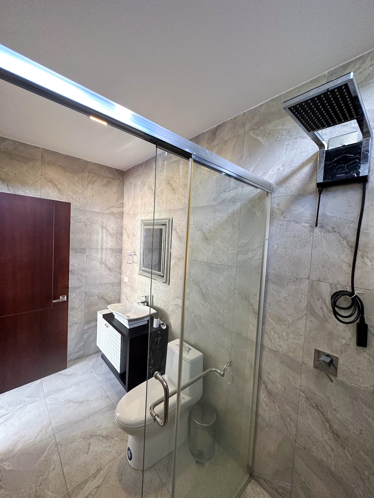 A modern bathroom features a glass shower enclosure with a rainfall showerhead. A sleek vanity with a wash basin is visible, alongside a toilet. The walls are adorned with light-colored tiles, and a dark wooden door is positioned in the background.