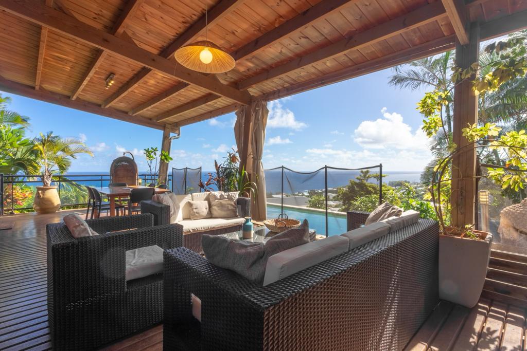 An outdoor seating area features a comfortable sectional sofa and a coffee table, set under a wooden pergola. Lush greenery surrounds the space, with views of the ocean visible in the background. A natural stone pool and safety netting are also part of this inviting setup.