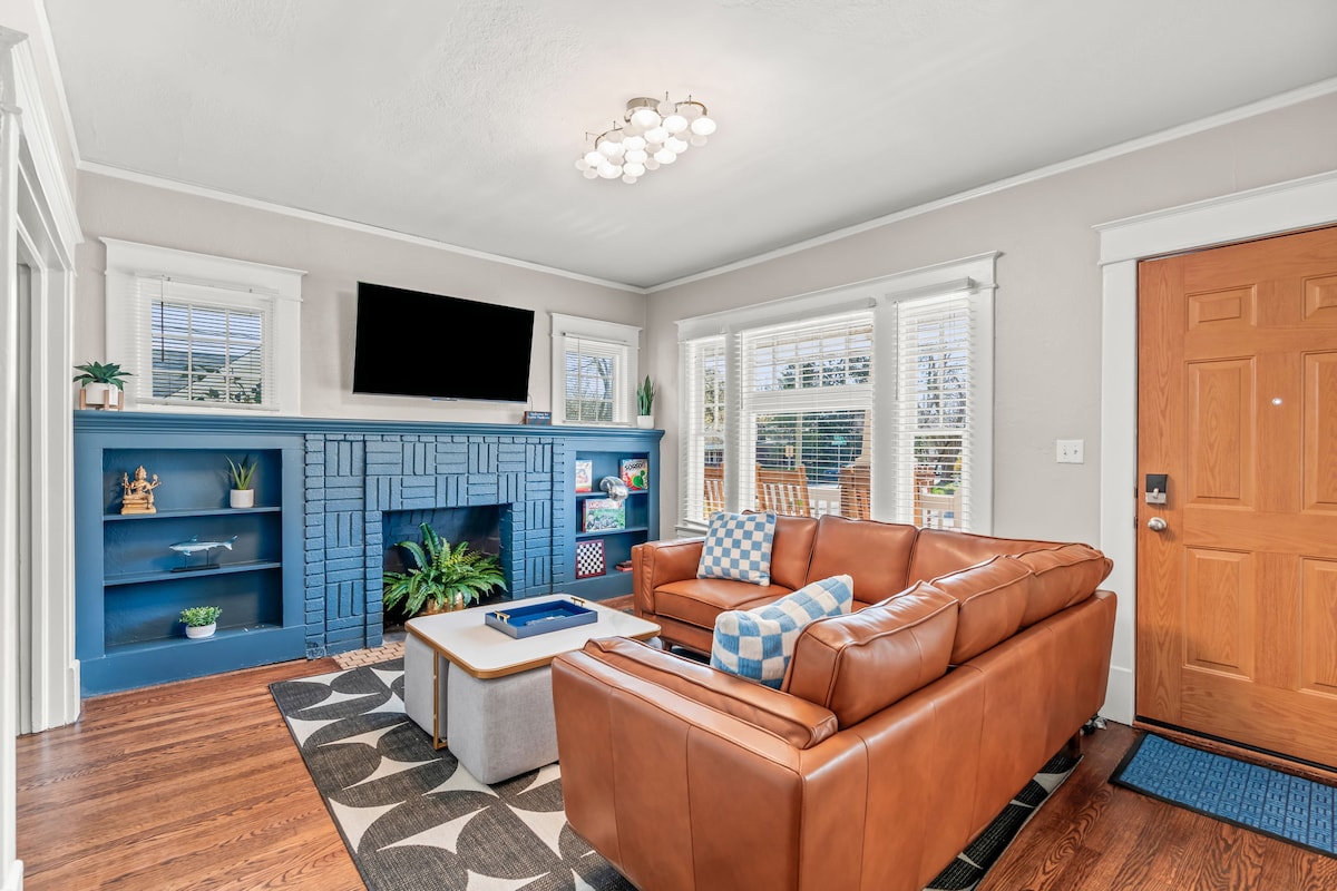 A living room with natural light streaming through multiple windows features a modern light fixture above a brown leather couch. A blue accent wall surrounds the fireplace, complementing a cozy rug and decorative shelves, creating a welcoming space for relaxation.