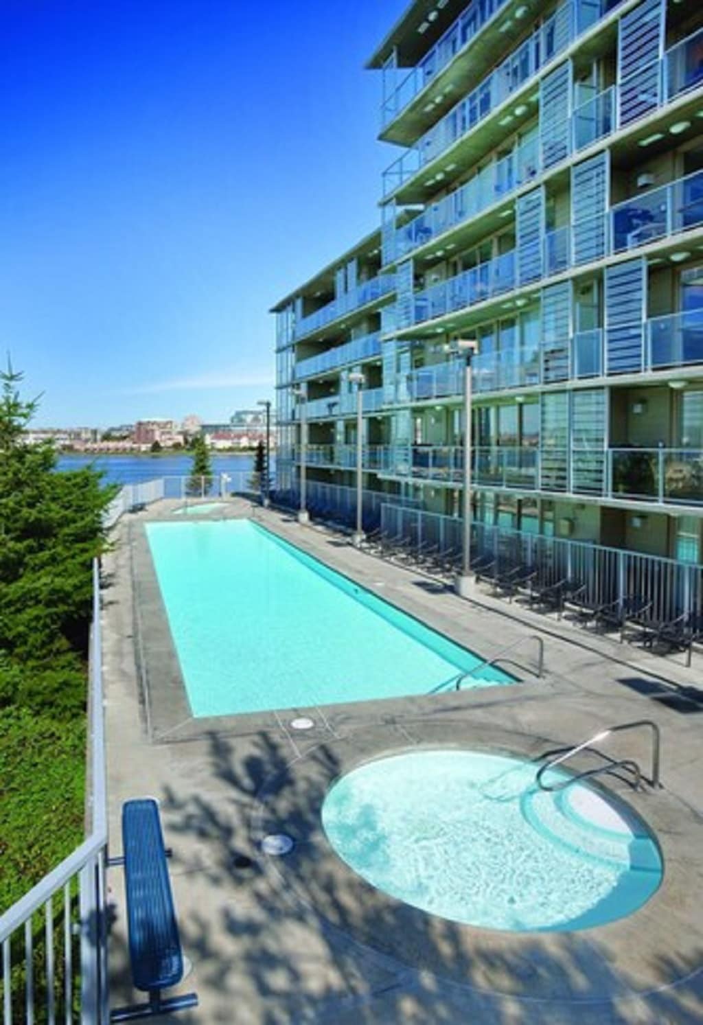 An outdoor swimming pool offers clear water alongside a hot tub, surrounded by a concrete deck with lounge chairs. The multi-story building, with a modern facade of glass and steel, provides a backdrop, while trees add greenery to the area.