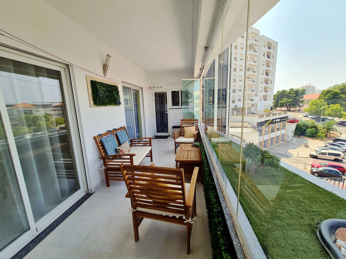 A private balcony is shown with comfortable wooden seating arranged around a small table, featuring a green lawn border. Large glass windows provide natural light and a view of the surrounding area, showcasing nearby buildings and greenery.