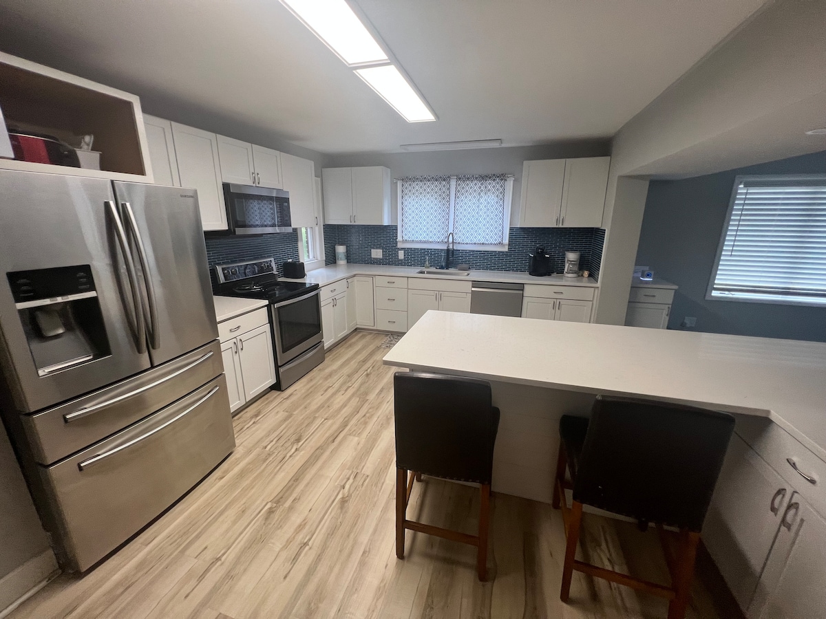 A modern kitchen is equipped with stainless steel appliances, including a refrigerator, oven, and microwave. Crisp white cabinets are complemented by a blue tile backsplash. A spacious island provides additional counter space and seating with two wooden stools. Natural light enters through a nearby window.