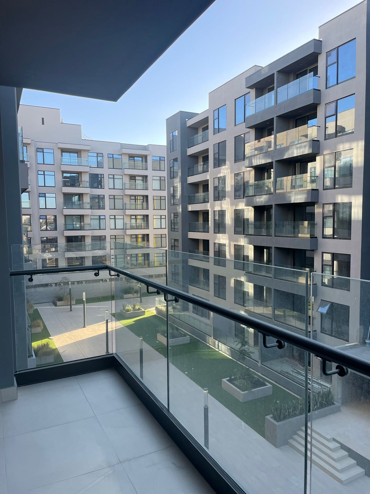 A balcony view reveals a modern apartment complex with multiple balconies and large windows. An open courtyard with landscaped greenery is visible below, featuring stone pathways and seating areas. The clear sky highlights the sleek architecture and contemporary design of the surrounding buildings.