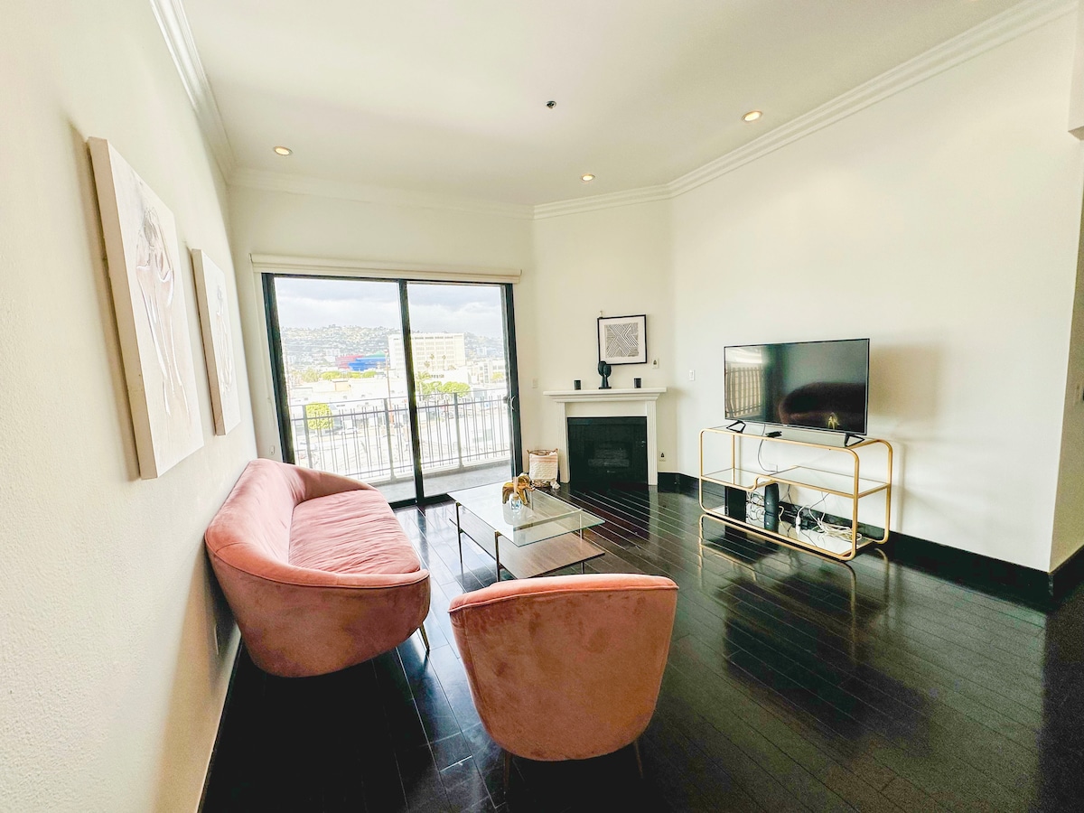 A modern living area features a pink curved sofa and a glass coffee table. Natural light fills the space through large sliding glass doors that lead to a balcony. A sleek television is mounted on a stand, while a decorative fireplace adds visual interest to the room.