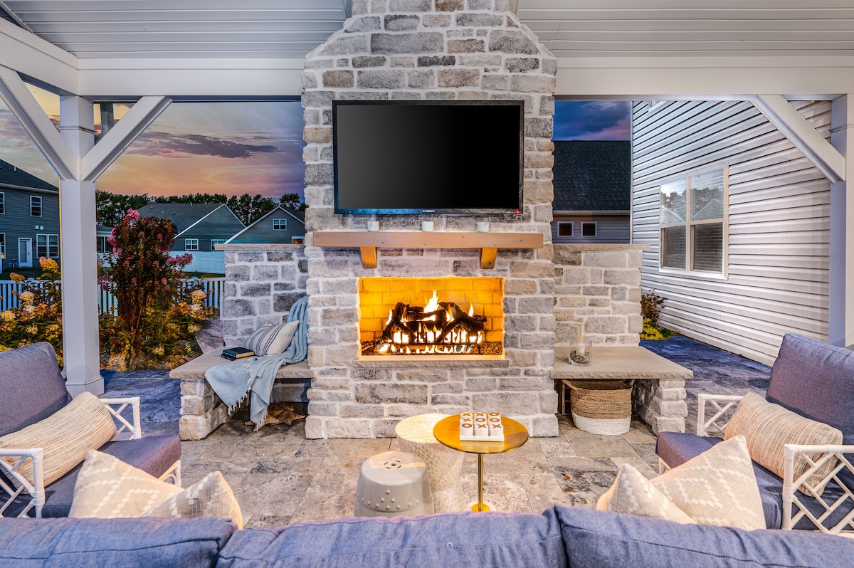 An outdoor living space is featured, showcasing a stone fireplace with a lit fire and a mounted TV above. Comfortable seating is arranged around a small table, with soft textiles adorning the cushions. The area is framed by wooden beams, offering a cozy yet open atmosphere.