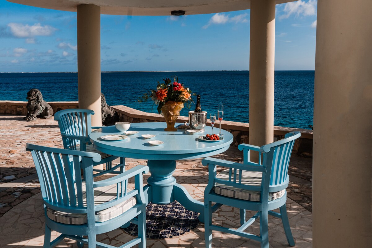 A circular blue table is set for dining with two decorative plates and a vase filled with flowers. Four matching chairs surround the table, with the ocean visible in the background, enhancing the peaceful atmosphere of the space.