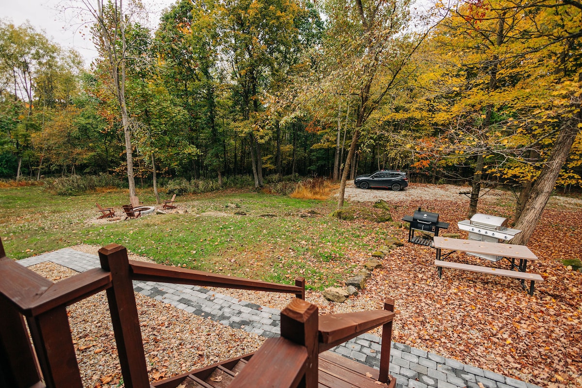 A peaceful outdoor scene is presented, showcasing a spacious yard surrounded by trees in autumn foliage. A gravel path leads to a barbecue grill and a picnic table, while a few seating areas with benches and fire pits are visible. A vehicle is parked nearby.
