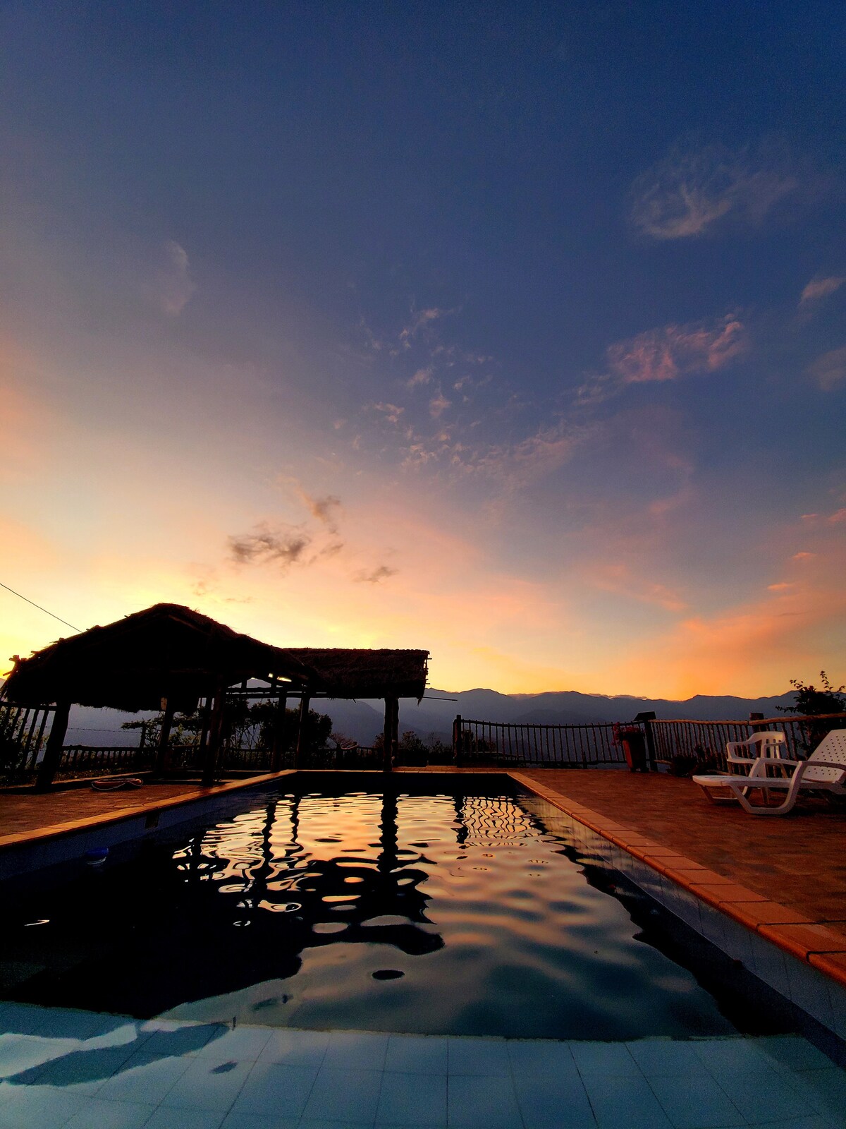 A tranquil outdoor pool reflects the colors of a dusk sky, with soft pink and orange hues blending into blue. A thatched-roof structure is visible nearby, providing shade. Two lounge chairs are positioned beside the pool, enhancing the peaceful ambiance of the setting.