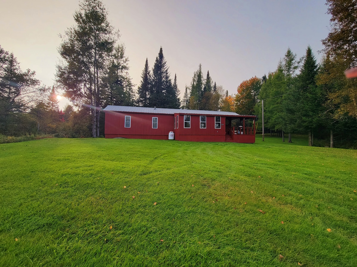A red wooden structure is set against a backdrop of lush green grass and dense trees. The late afternoon sun casts a warm light, enhancing the vibrant colors of the natural landscape. A deck extends from the building, offering an outdoor space for relaxation.