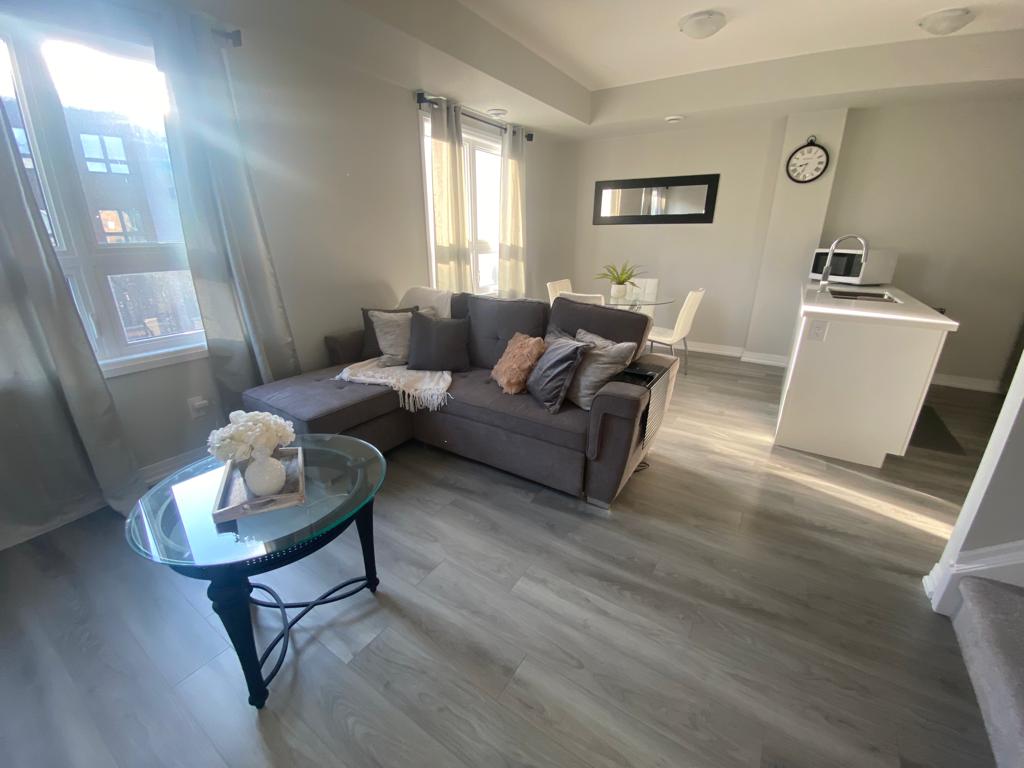 A modern living space features a comfortable gray couch adorned with decorative pillows. Light streams through large windows, illuminating the wooden floor. A round glass coffee table sits in front of the couch, while a dining area with a white table and chairs is visible in the background.