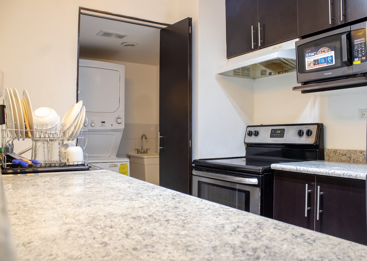 The kitchen area features dark cabinetry and a spacious countertop. A stove and microwave are present alongside a sink area. An equipped laundry area with a washing machine and dryer is visible in the background, separated by a door.