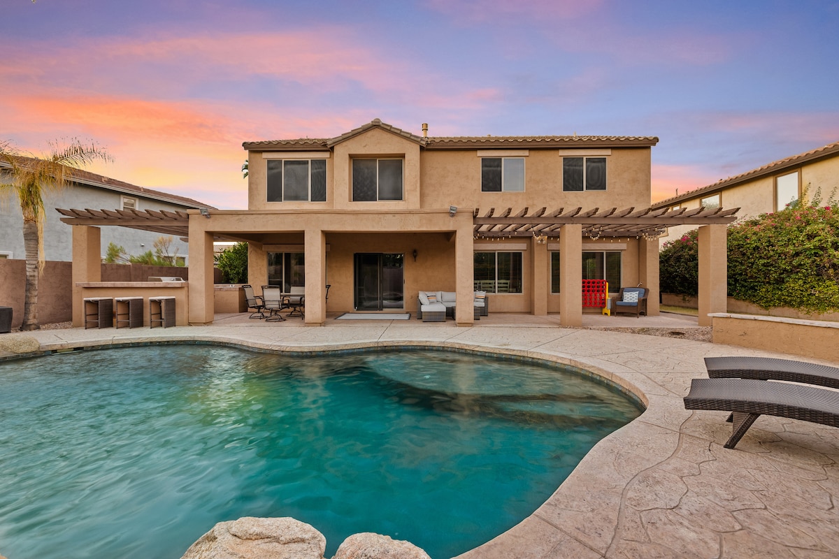 A two-story home is depicted with an expansive outdoor area featuring a shimmering pool. The patio is shaded by wooden pergolas, and seating arrangements are seen positioned around the pool, creating an inviting space for relaxation. Colorful landscaping adds a natural touch.