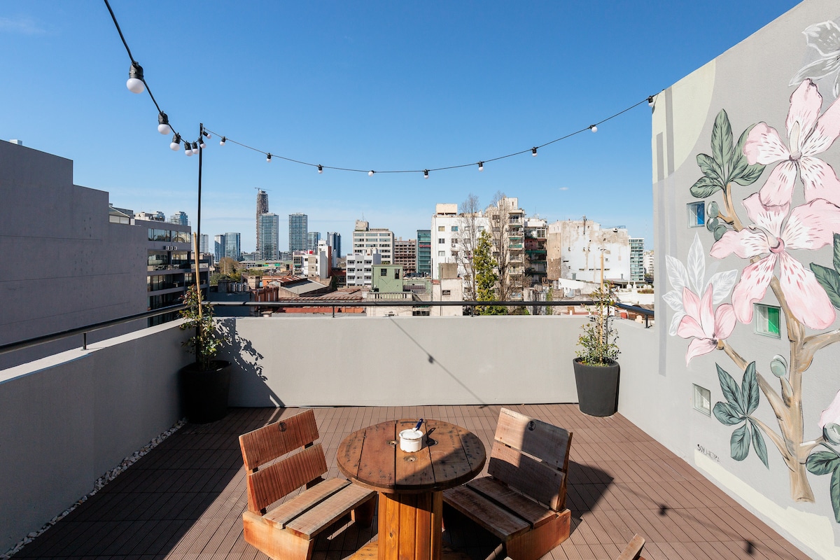 A private terrace is showcased, featuring a round wooden table surrounded by two wooden chairs. String lights are arranged above, enhancing the outdoor space. In the background, city buildings rise against a clear blue sky, with a mural of pink flowers adorning one wall.