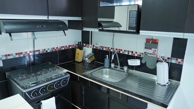 A modern kitchen is equipped with a gas stove, oven, and a microwave above for efficient cooking. The sink is positioned next to the countertop, adorned with utensils and a dish rack. Colorful tile accents enhance the sleek black cabinetry.
