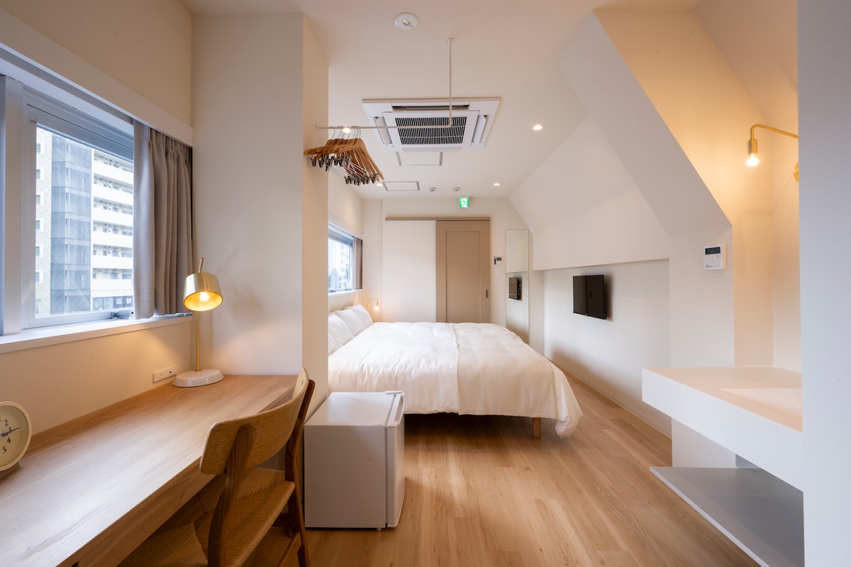 A spacious bedroom features two beds with crisp white bedding, complemented by soft natural lighting from two large windows. A wooden desk and two chairs are arranged alongside a small refrigerator, creating a functional area. Wall-mounted televisions are positioned for easy viewing.