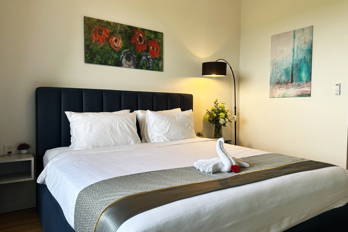 A spacious room is presented with a large bed featuring white linens and decorative pillows. Above the bed, two pieces of artwork add color, while a floor lamp and a small table are positioned nearby. Soft natural light enters through a window, enhancing the serene ambiance.