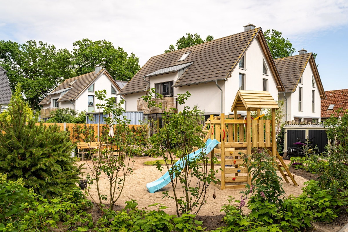 A charming outdoor play area is presented, featuring a sandbox surrounded by lush greenery. A slide and climbing structure are positioned on the soft sand, with colorful landscaping and nearby trees providing shade. The house's exterior stands in the background, showcasing a pleasant residential setting.