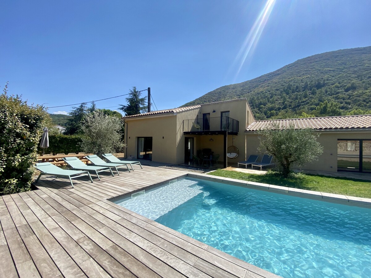The exterior of a modern villa is showcased, featuring a clear blue pool surrounded by wooden decking. Lounge chairs are arranged nearby, with olive trees adding greenery. The hillside in the background creates a serene backdrop under a bright blue sky.